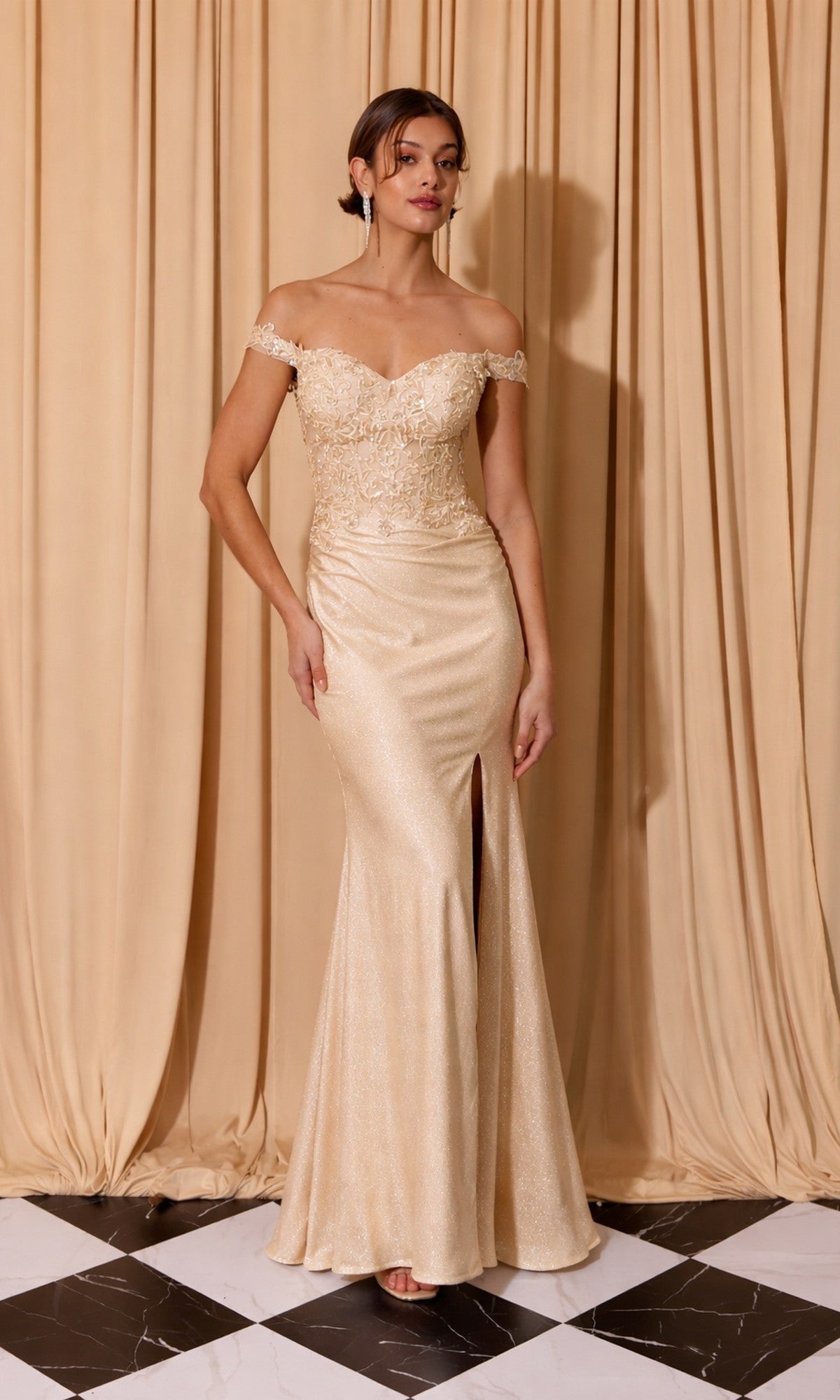 A woman stands on a black and white tiled floor in front of beige curtains, wearing the Eureka EK173 long prom dress in champagne, featuring an off-shoulder floral embroidered corset and front slit.